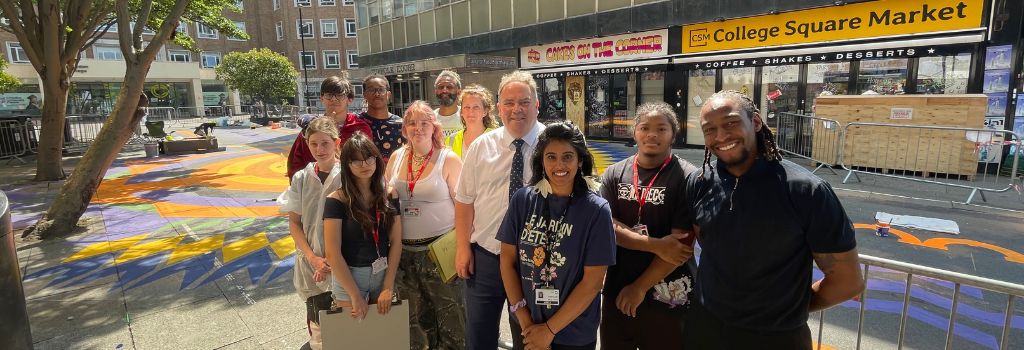 Mayor Perry stands with students and artists at the new Croydon Compass artwork in Croydon Square. It is very sunny.